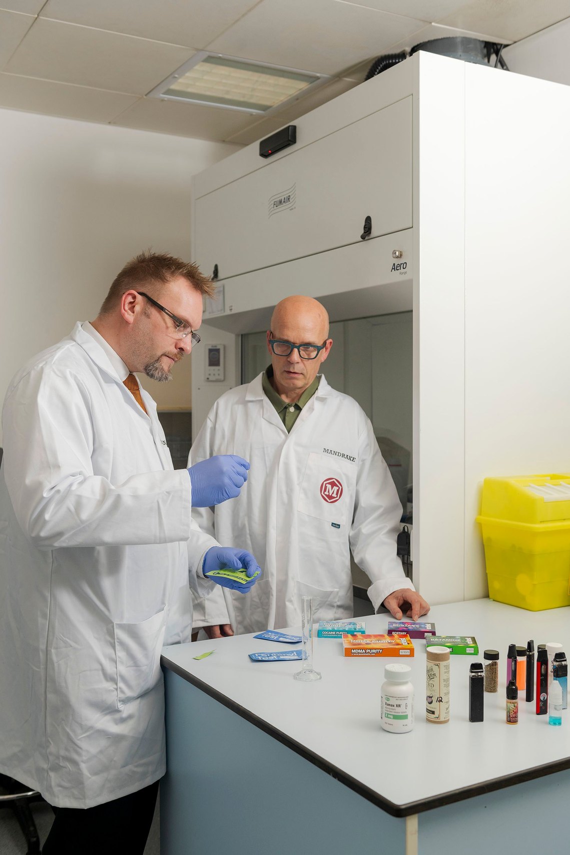 Two scientists in lab coats stand at a lab bench with various samples and equipment. Brand and marketing photography crafted by a Manchester freelance photographer.