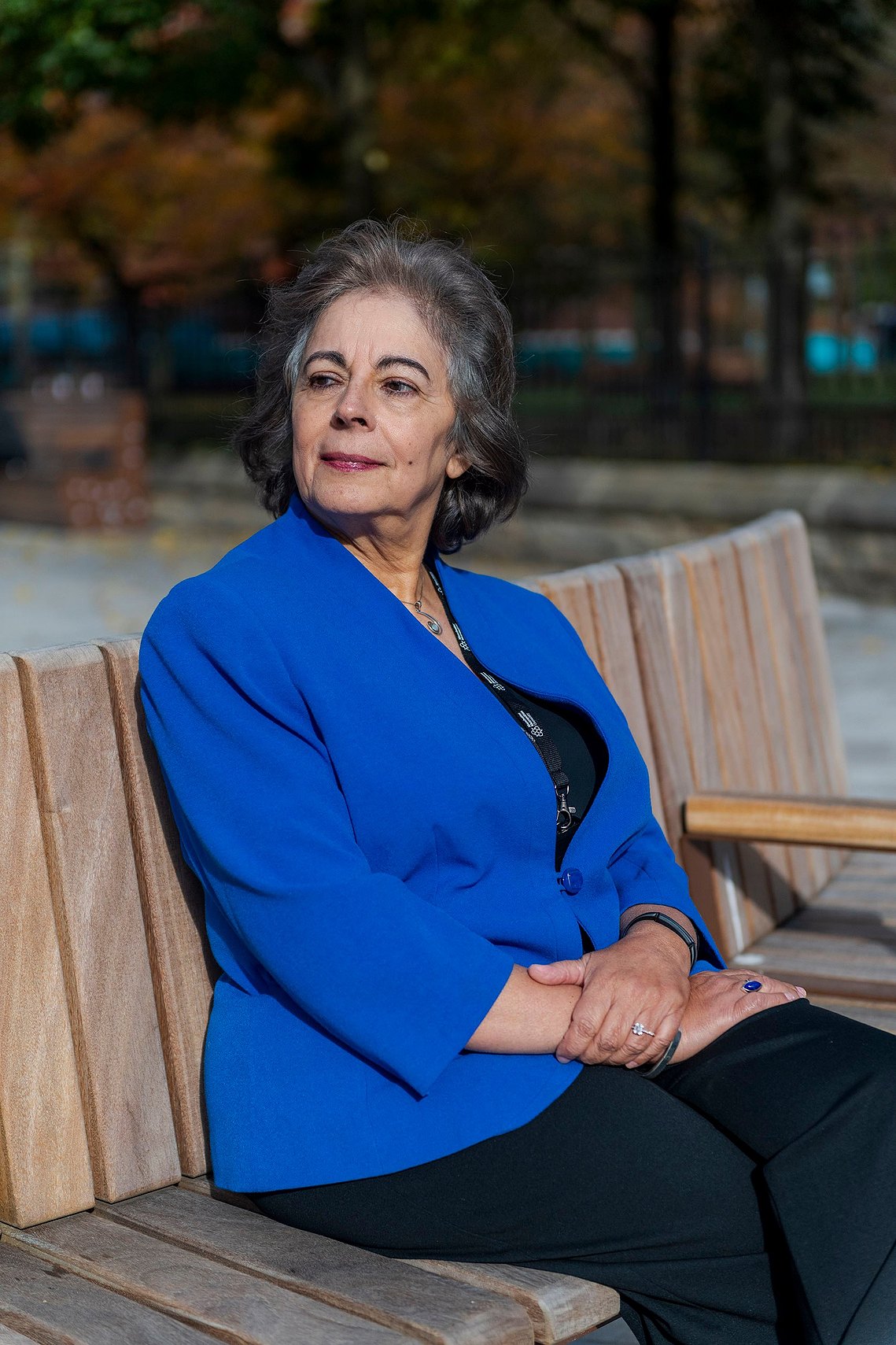 An woman wearing a bright blue jacket sits on a wooden bench with a thoughtful expression. Trees are blurred in the background.
