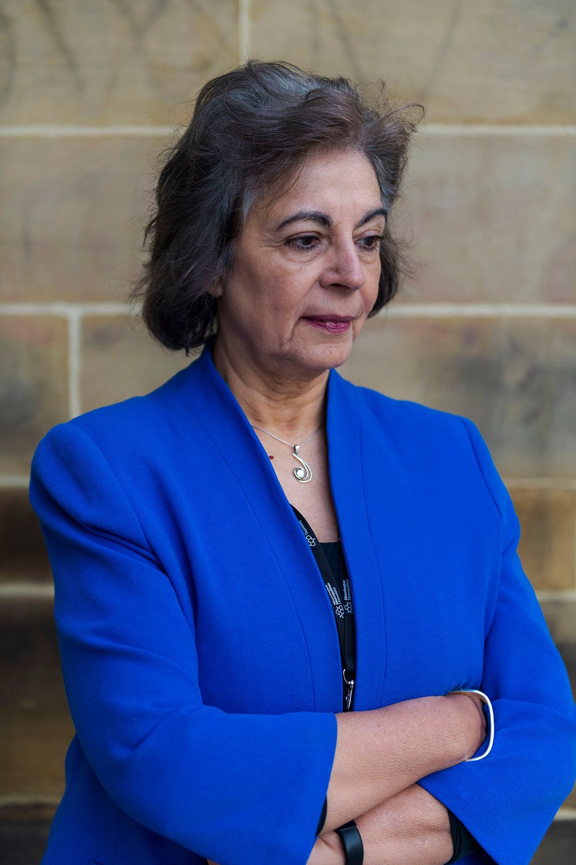 A woman with her arms crossed, wearing a bright blue blazer over a dark top. She is gazing downward against a background of light brown stone tiles.