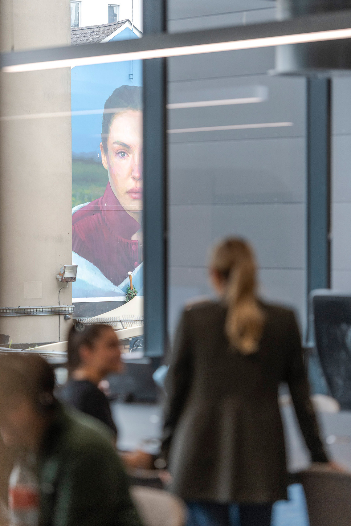 Authentic lifestyle photography of members working naturally at Clockwise workspace. Subjects out of focus in foreground and large Belfast mural visible outside
