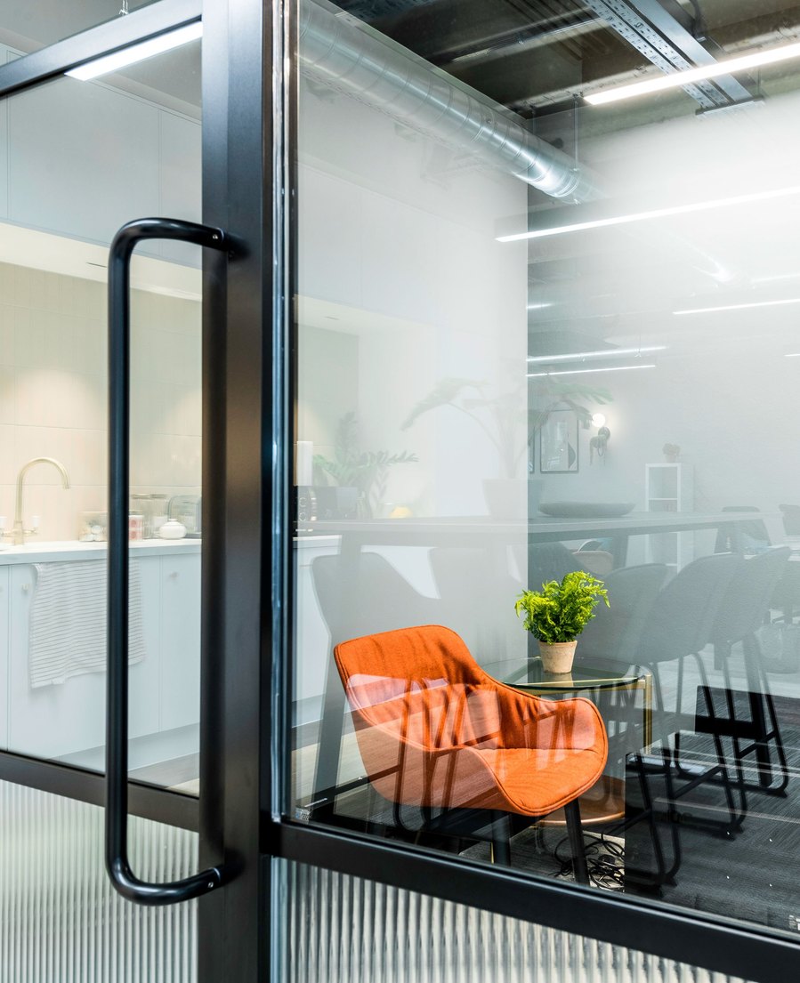 Modern office lounge viewed through glass door with an orange chair and a plant on the table.