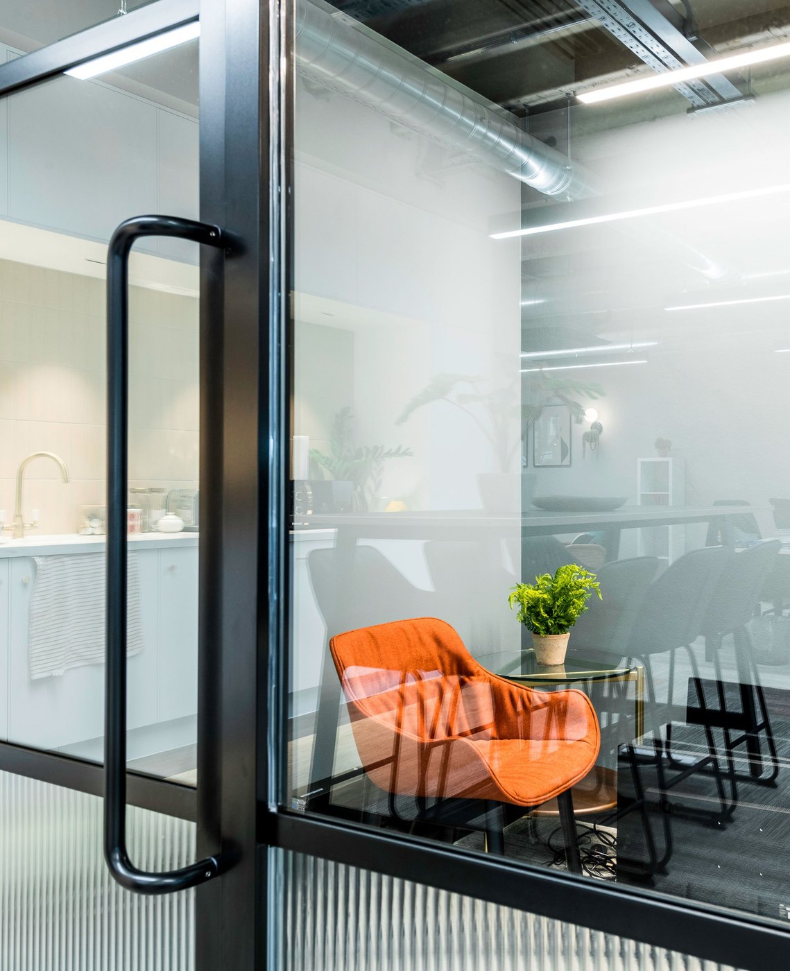 Modern office interior viewed through glass door, featuring an orange chair and potted plant.