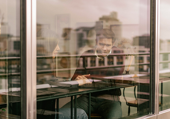 Commercial lifestyle photography showing workers chatting over a laptop at a window seat. Image taken from outside the window
