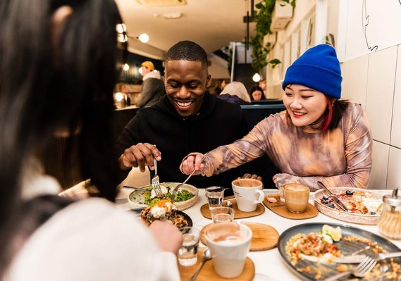 Two young people in a restaurant smiling as they tuck into shared sites of food.