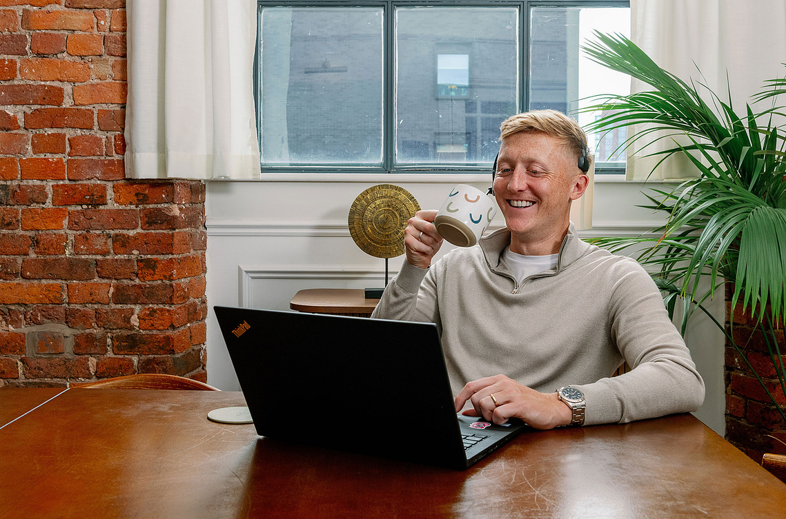 Professional yet relaxed brand photography Manchester. Man drinks from cup and smiles at laptop. Brickwork and plants in background.
