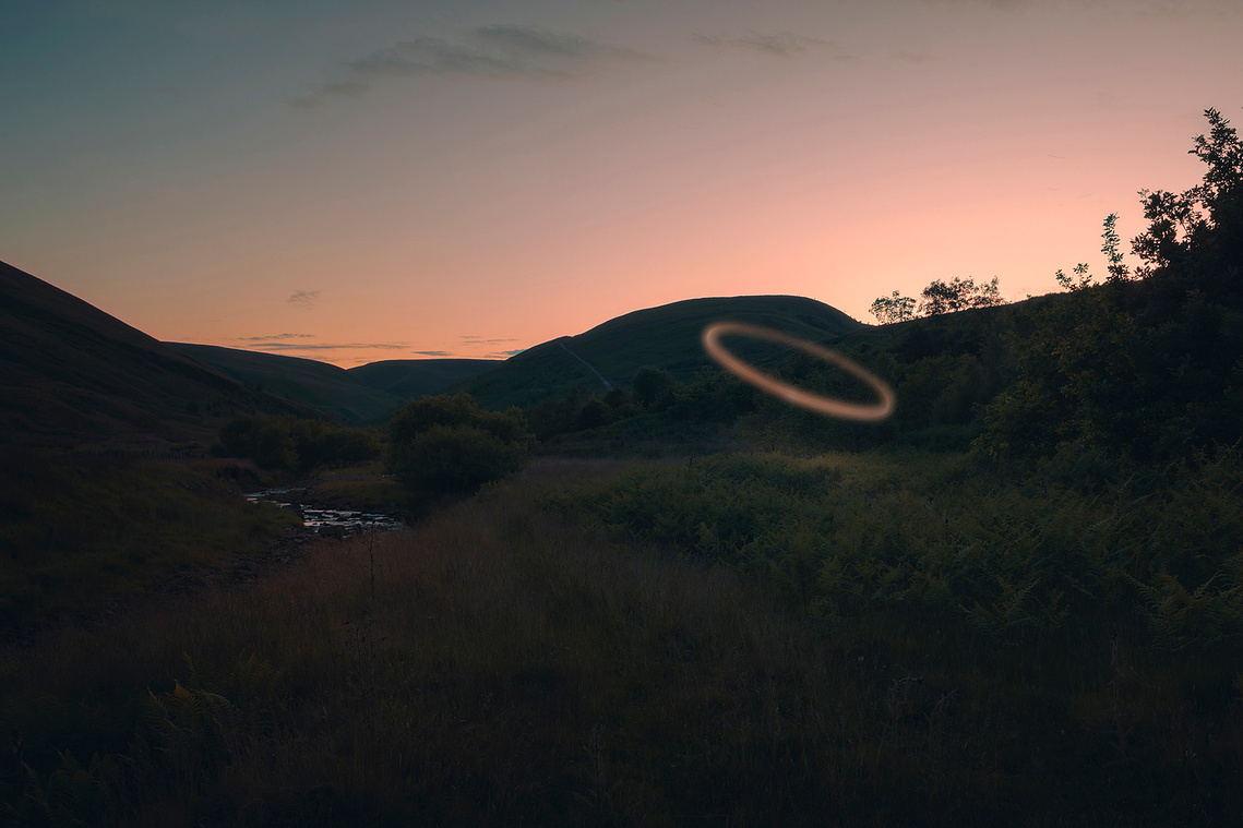 a ring of light dominates this landscape photograph taken at twilight, dark foreground, a small stream visible and the last of the sunset just visible behind the hills. 