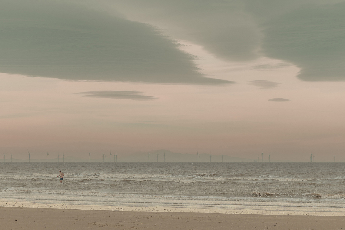 beach late afternoon, choppy waves, wind turbines visible in background, swimmer emerges from water in red hat and swimmers
