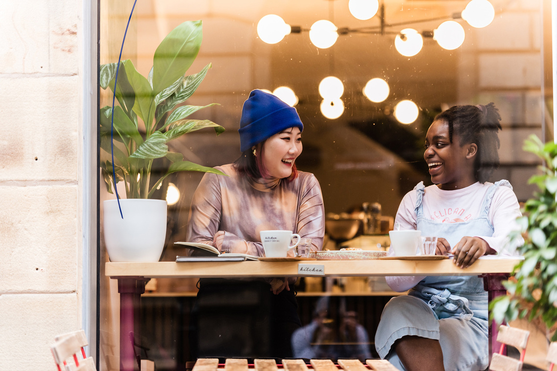 Two young students sat in window of coffee shop enjoying a hot drink and laughing 