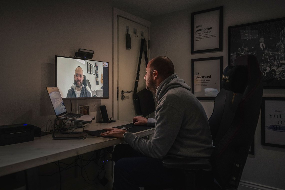 person in home office, basement office with no obvious daylight, man has home office setup with laptop and screen and seems to be on a conference call with a colleague