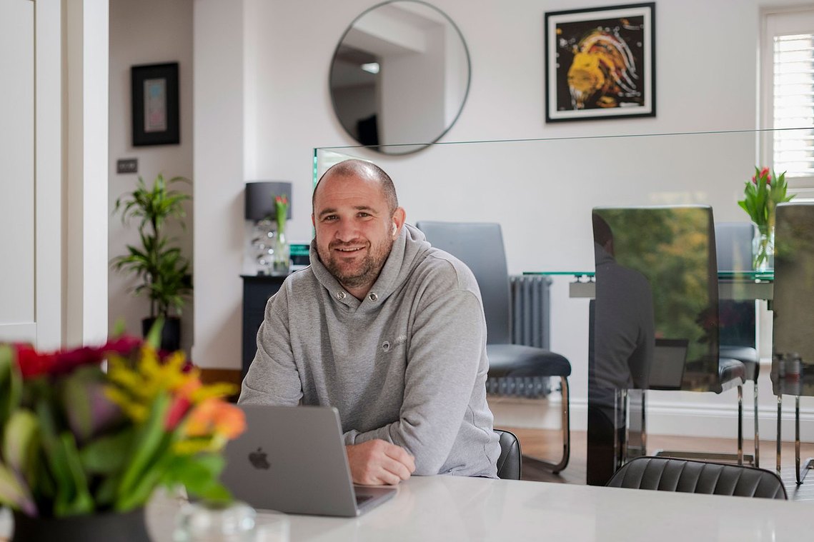 person working from home, open plan kitchen environment with pictures on wall and flowers in background. man smiles at camera with laptop in front of him