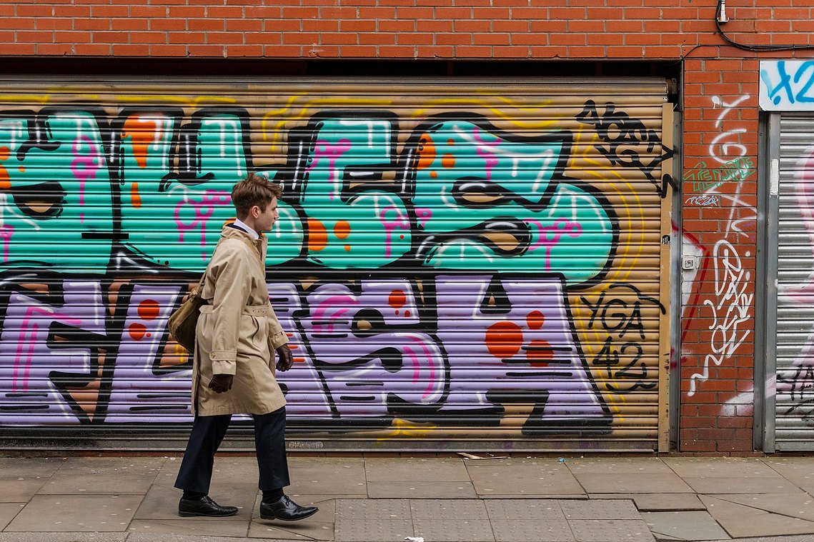 architecture and lifestyle photography, manchester street photography, young man well dressed in rain mac and gloves, walks past graffiti in urban scene