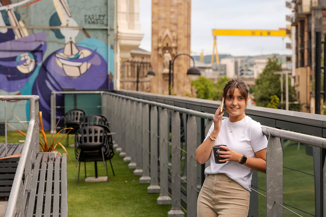 Natural light workspace photography showing people enjoying exterior breakout areas. Girl on phone with mug in hand. Belfast skyline visible behind.
