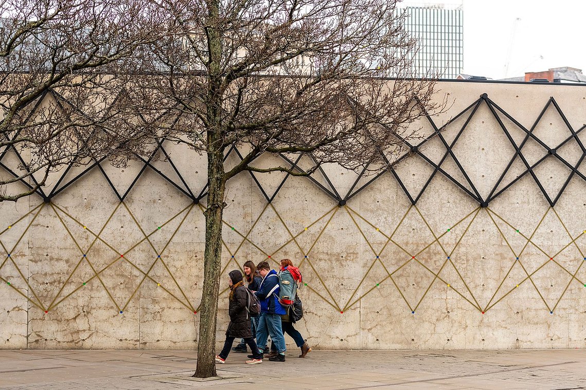 architecture and lifestyle photography, manchester street photography, group of people walk past large wall with geometric pattern on it. urban scene with a tree