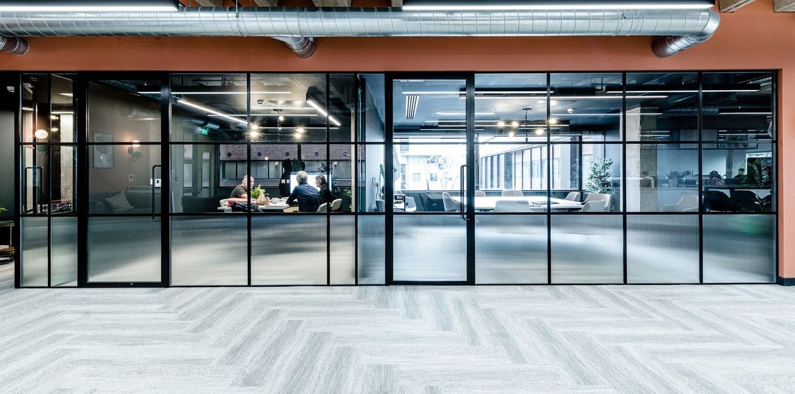 Modern office space with glass walls, people working at tables, and exposed ductwork overhead. commercial interiors photography