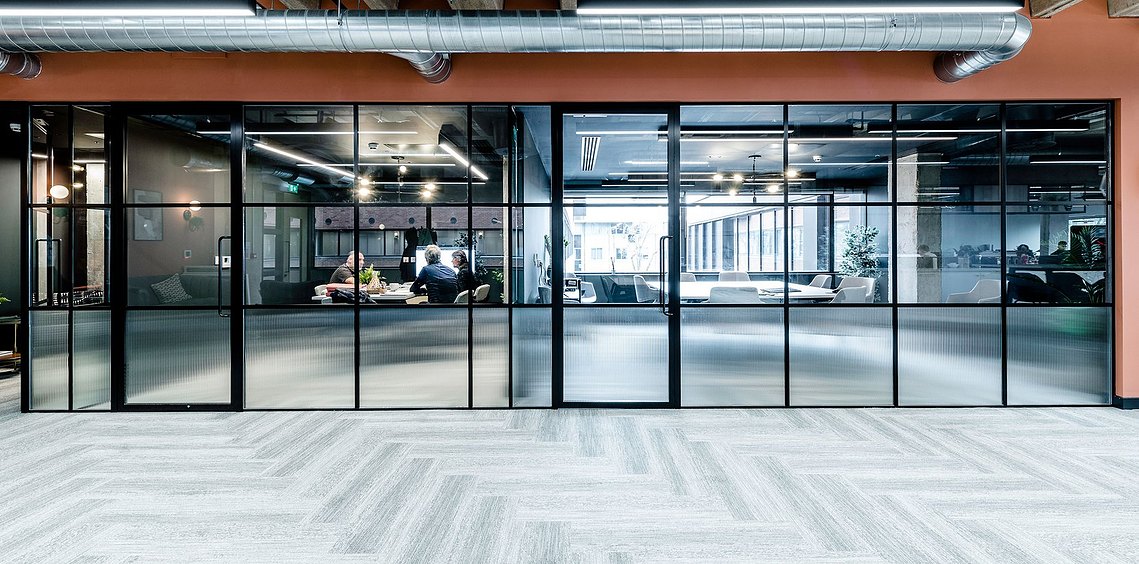 Modern office space with glass walls, people working at tables, and exposed ductwork overhead. commercial interiors photography
