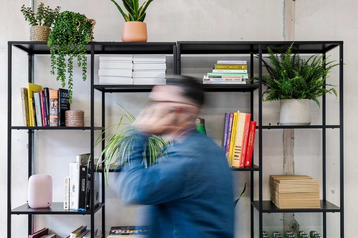 commercial lifestyle photography showing staff in a workplace, staff member walks past a bookshelf with creative themed books while on phone