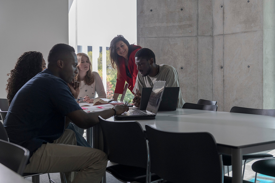 students and tutor grouped around a desk with paper and pens on desk and laptops, group are interacting and discussing project 