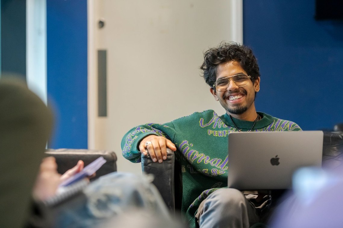 Person smiling while sitting on a chair with a laptop, wearing glasses and a green sweater.