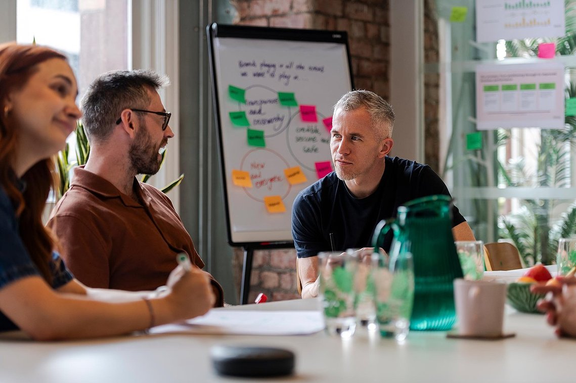commercial lifestyle photography showing staff in a workplace, team members sat at table discussing work, with plants and natural light