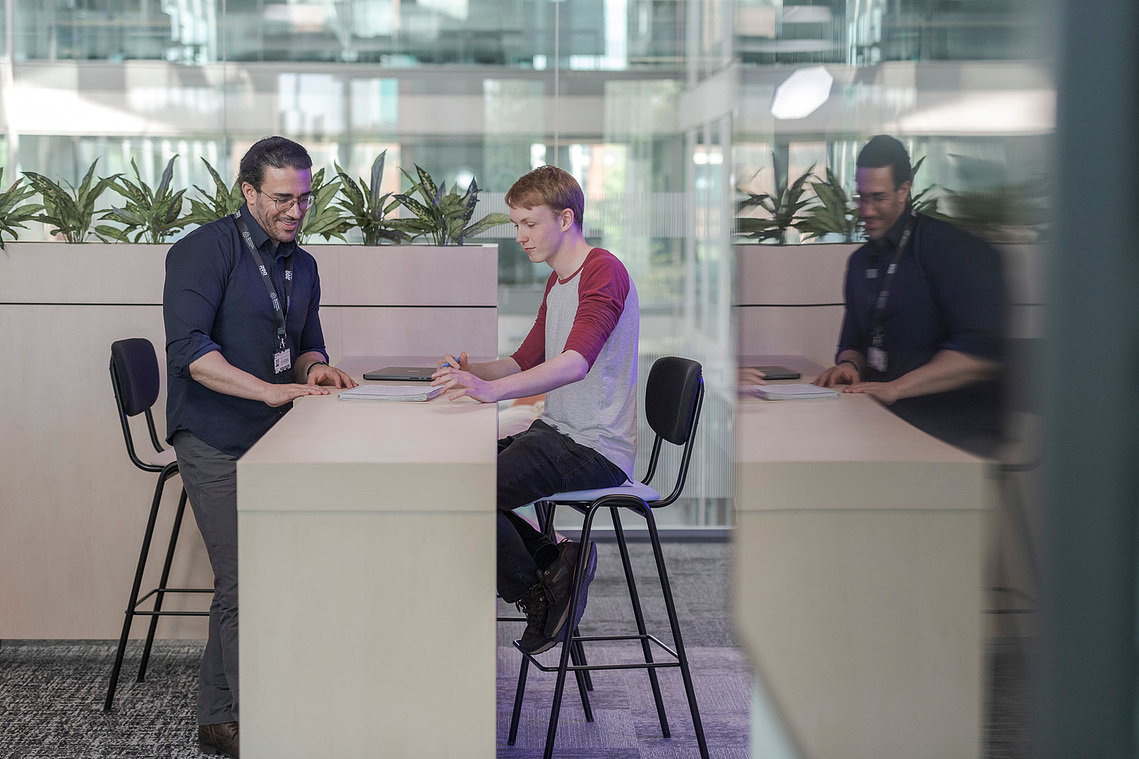 student and tutor discussing work at a counter with chairs, reflection in glass, both smiling 