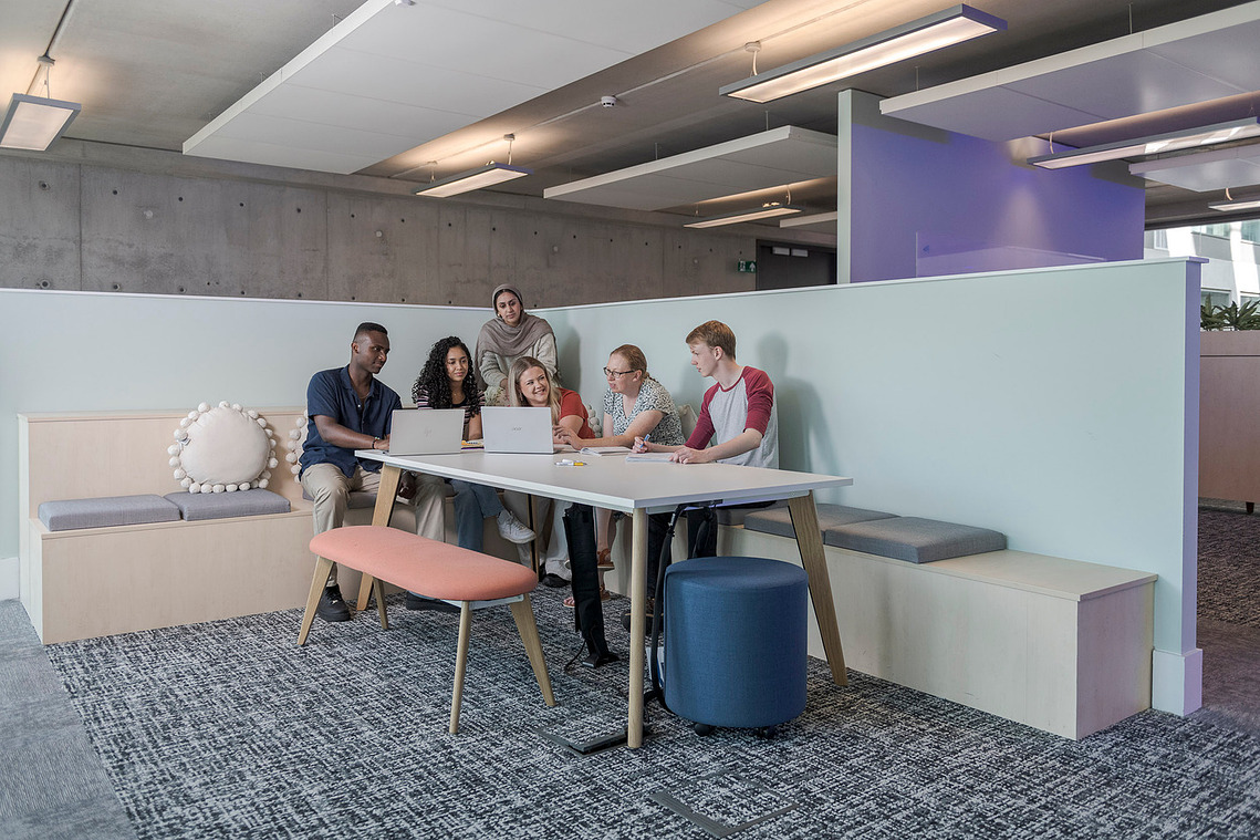students grouped around a desk in a modern university setting, group are discussing work on a laptop in front of them