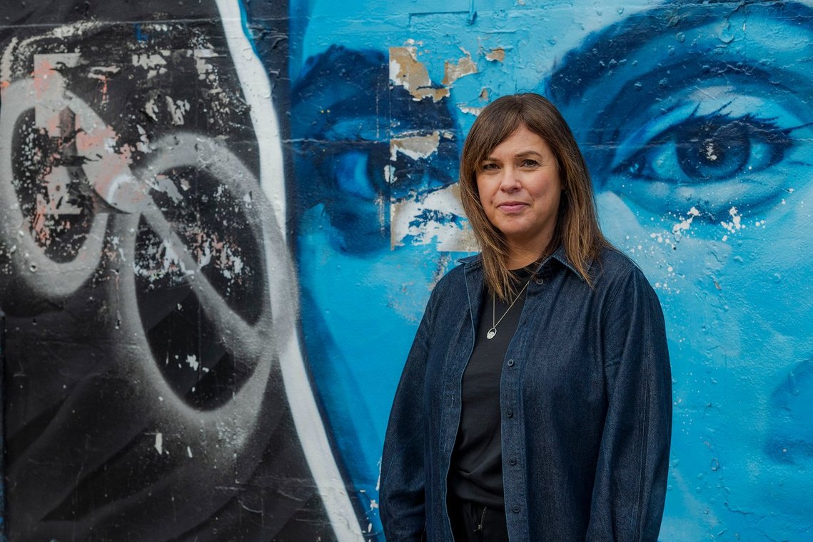 A woman stands in front of graffiti. She wears a dark denim shirt and black top, appearing thoughtful. Captured by a Manchester freelance photographer.