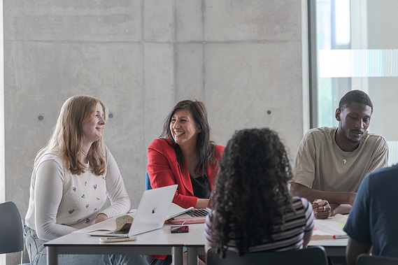 tutor sits with students smiling as they work on a project using laptops and notes