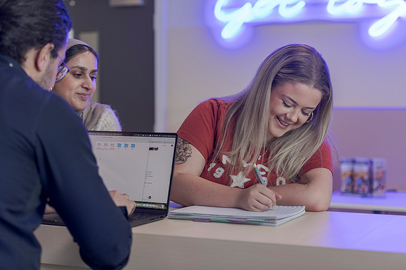 student writing and smiling as she discusses work with tutor who is working on a laptop, neon sign in background
