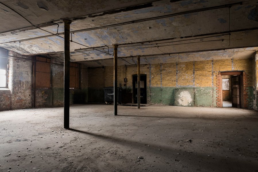 Abandoned industrial room with peeling walls, support columns, and a solitary garbage bin.