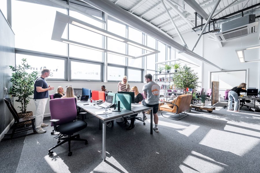Modern office space with people working at desks near large windows.