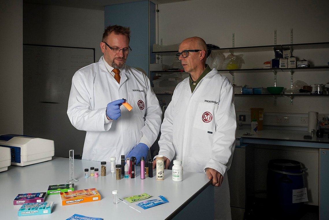 Two scientists in lab coats stand at a lab bench with various samples and equipment. Brand and marketing photography crafted by a Manchester freelance photographer.