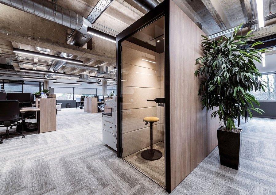 Modern office space with desks, chairs, exposed ductwork, and a large potted plant.