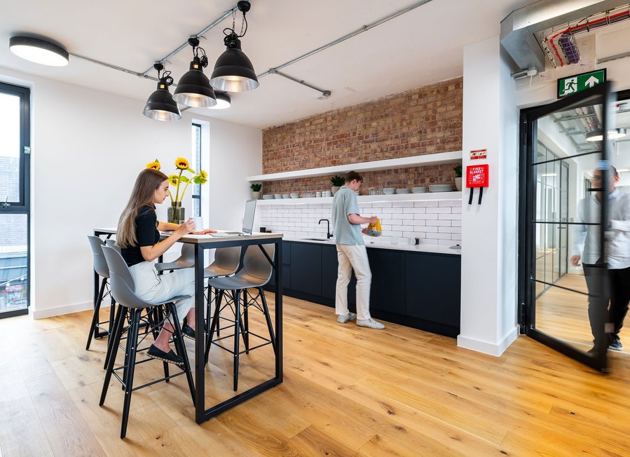 Modern office kitchenette with people, sunflowers on table, exposed brick wall, and industrial lights.