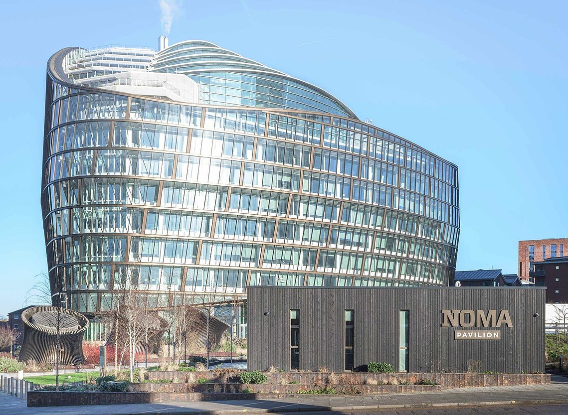 foreground shows single storey wooden clad building with large coop glass modern building in background