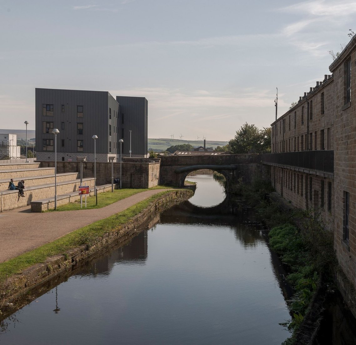 Exterior of Root Fifty-Two's office in Burnley, showing a row of stone terraced buildings with a canal in foreground. In far background wind turbines are visible on rolling hills.