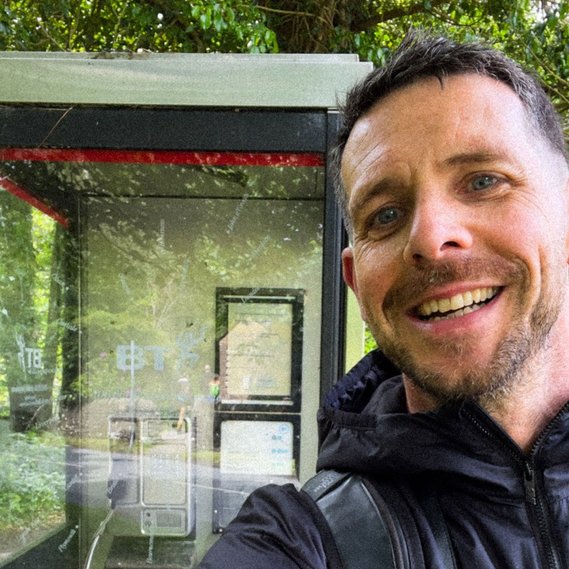 selfie of the artist in front of a phone box with trees behind.
