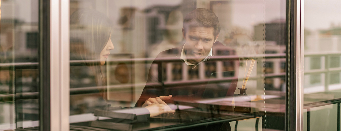 office lifestyle photography, workers consult over laptop, taken through window 