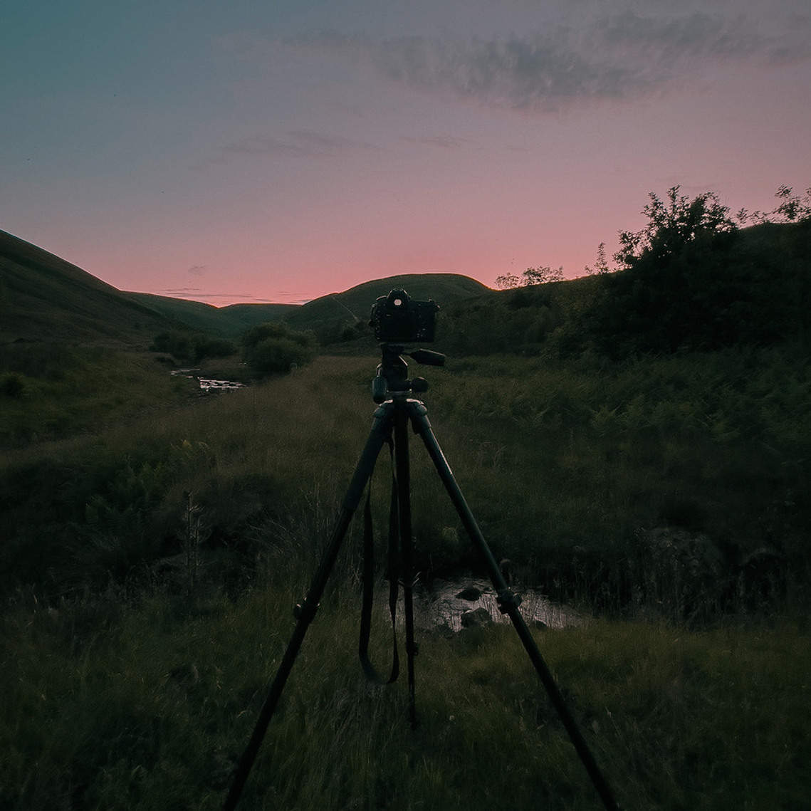 a landscape at twilight, camera on tripod in foreground, a small stream visible and the last of the sunset just visible behind the hills. a ring of light dominates the image.