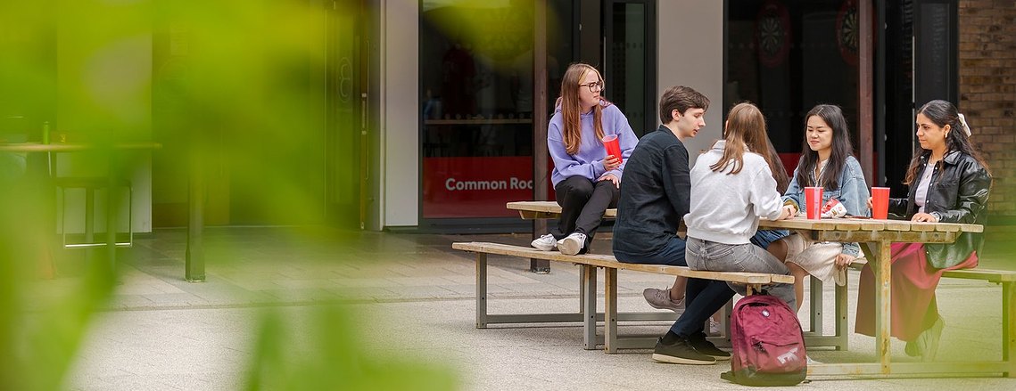 Five student sit at an outdoor table, talking and holding drinks, capturing the essence of student lifestyle photography. A building stands in the background, framed by lush greenery in the foreground