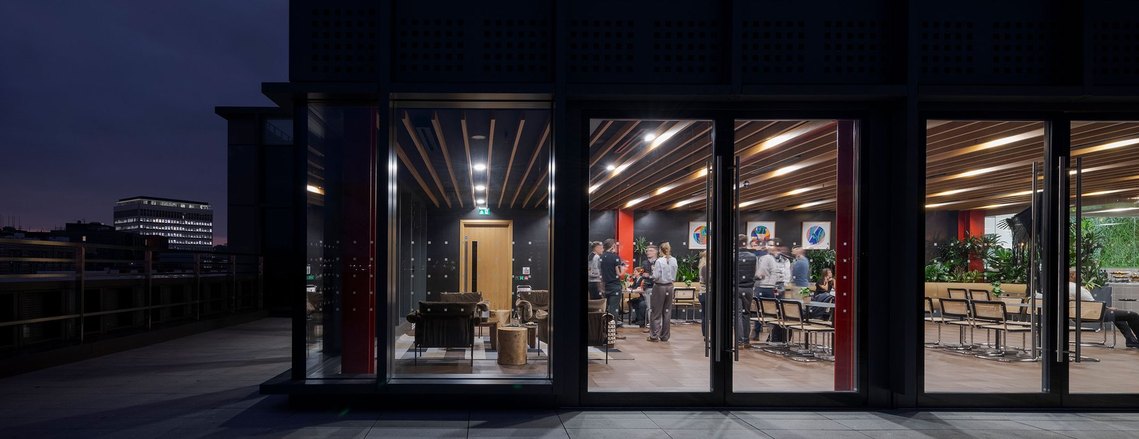 architecture photography of social space on top floor of a building, with a terrace outside. people visible inside and view of bristol at dusk in background