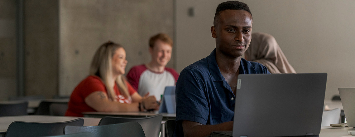 student sat at desk looking at laptop, other students in background, class environment, university, higher education 