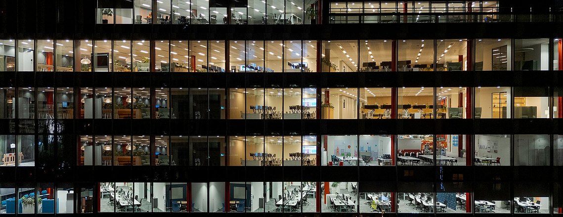 side on shot of a building lit up at night, taken from a drone, offices and furniture visible
