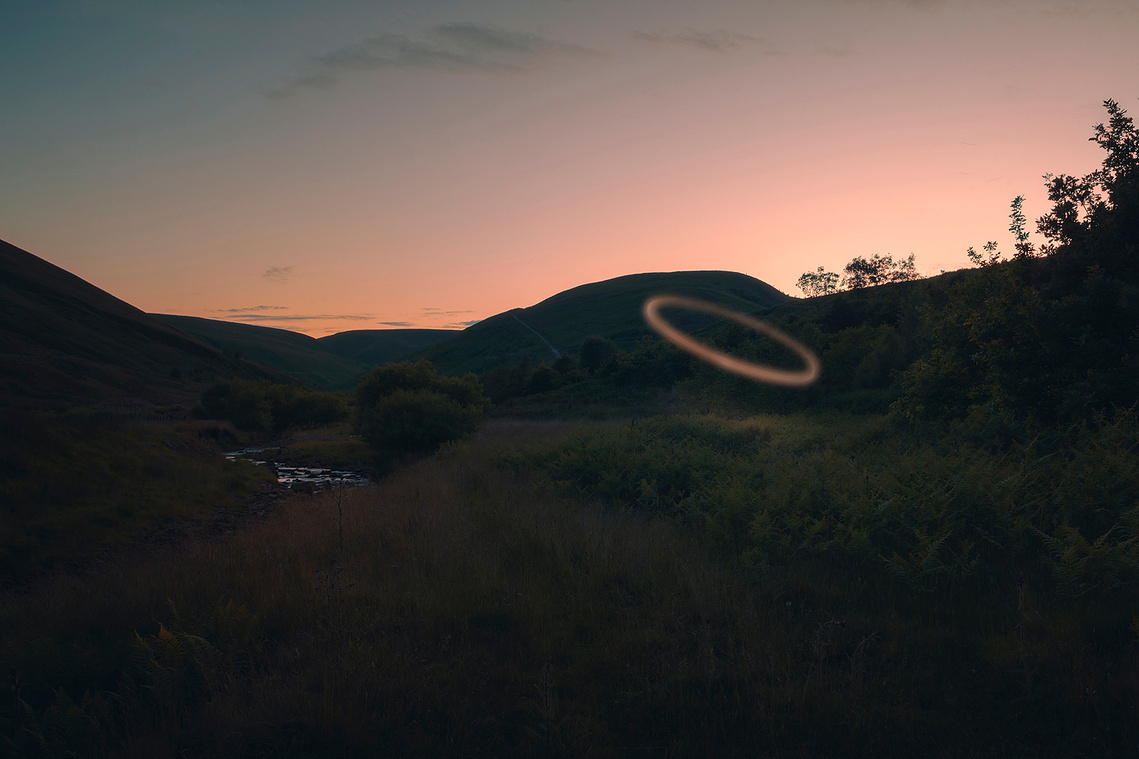 a landscape at twilight, dark foreground, a small stream visible and the last of the sunset just visible behind the hills. a ring of light dominates the image.