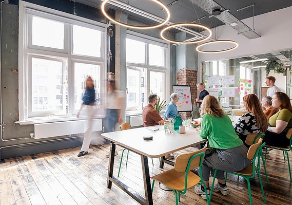 Commercial lifestyle photography shows a workplace group meeting with workers sat around table and walking around