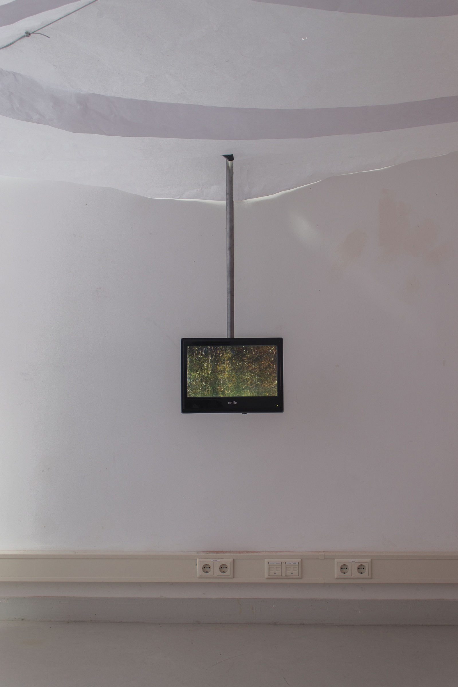 A room with paper ceiling. There is a video monitor on to behind coming down from the ceiling. On the monitor is a blurry image of green foliage. Jamie Kane