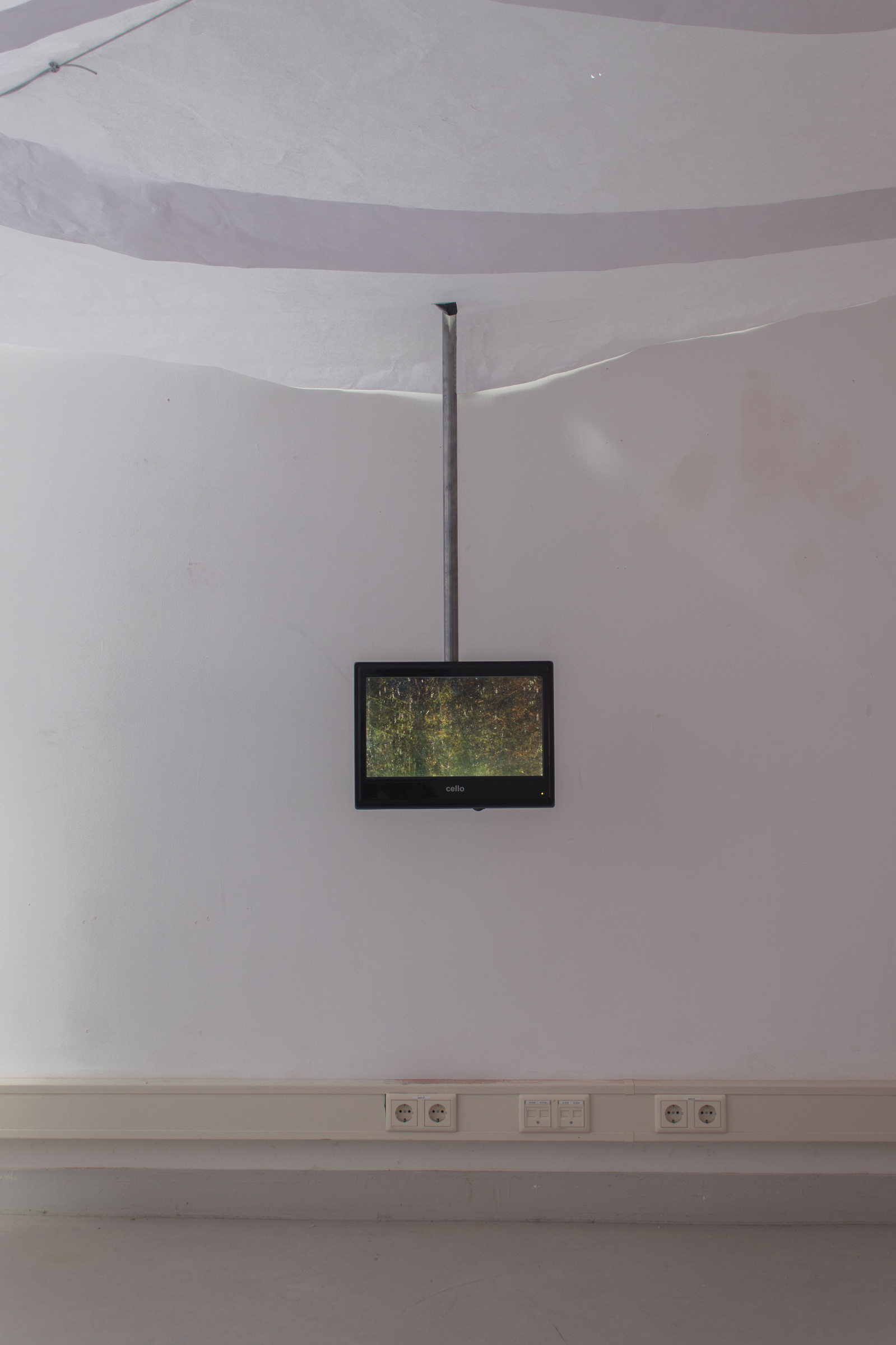 A room with paper ceiling. There is a video monitor on to behind coming down from the ceiling. On the monitor is a blurry image of green foliage. Jamie Kane