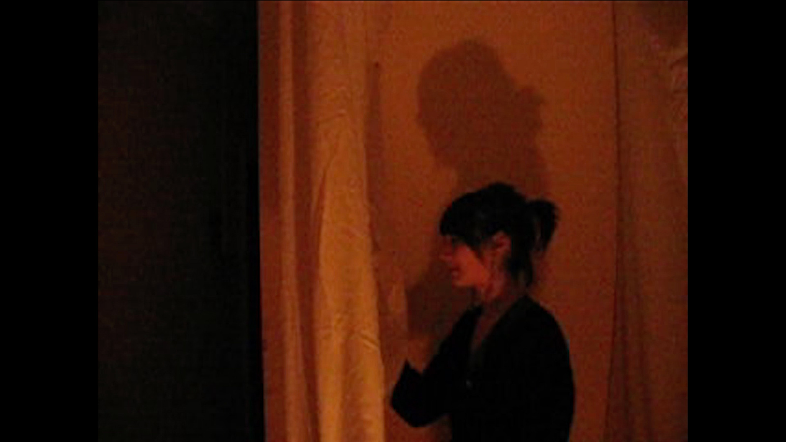 A woman with her shadow behind pulling open a curtain under orange light. Jamie Kane