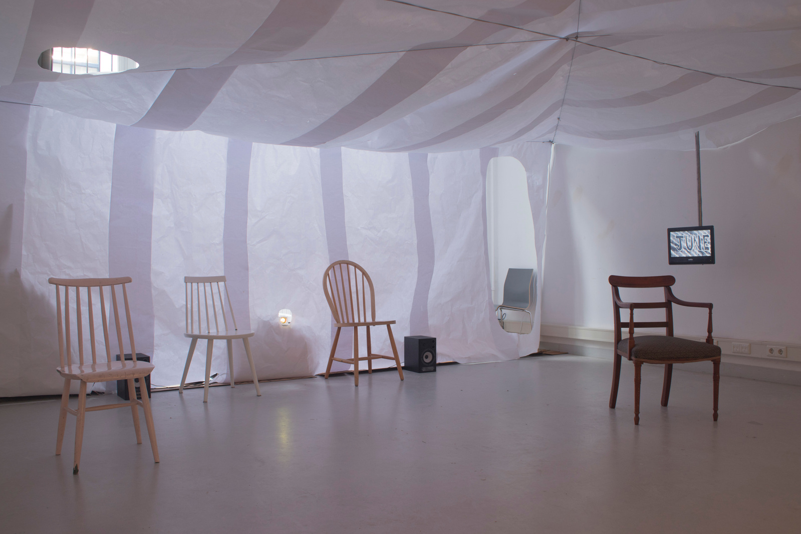 A room with paper ceiling and walls filled with wooden domestic chairs. There is a video monitor on to right coming down from the ceiling. Jamie Kane