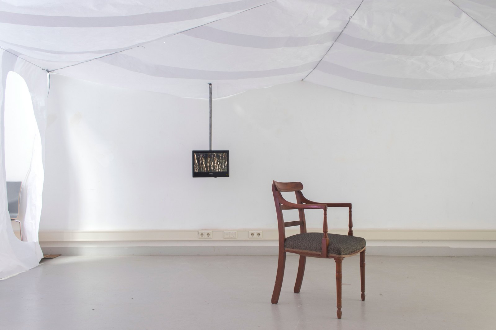 A room with paper ceiling and walls a wooden domestic chair. There is a video monitor on to behind coming down from the ceiling. Jamie Kane