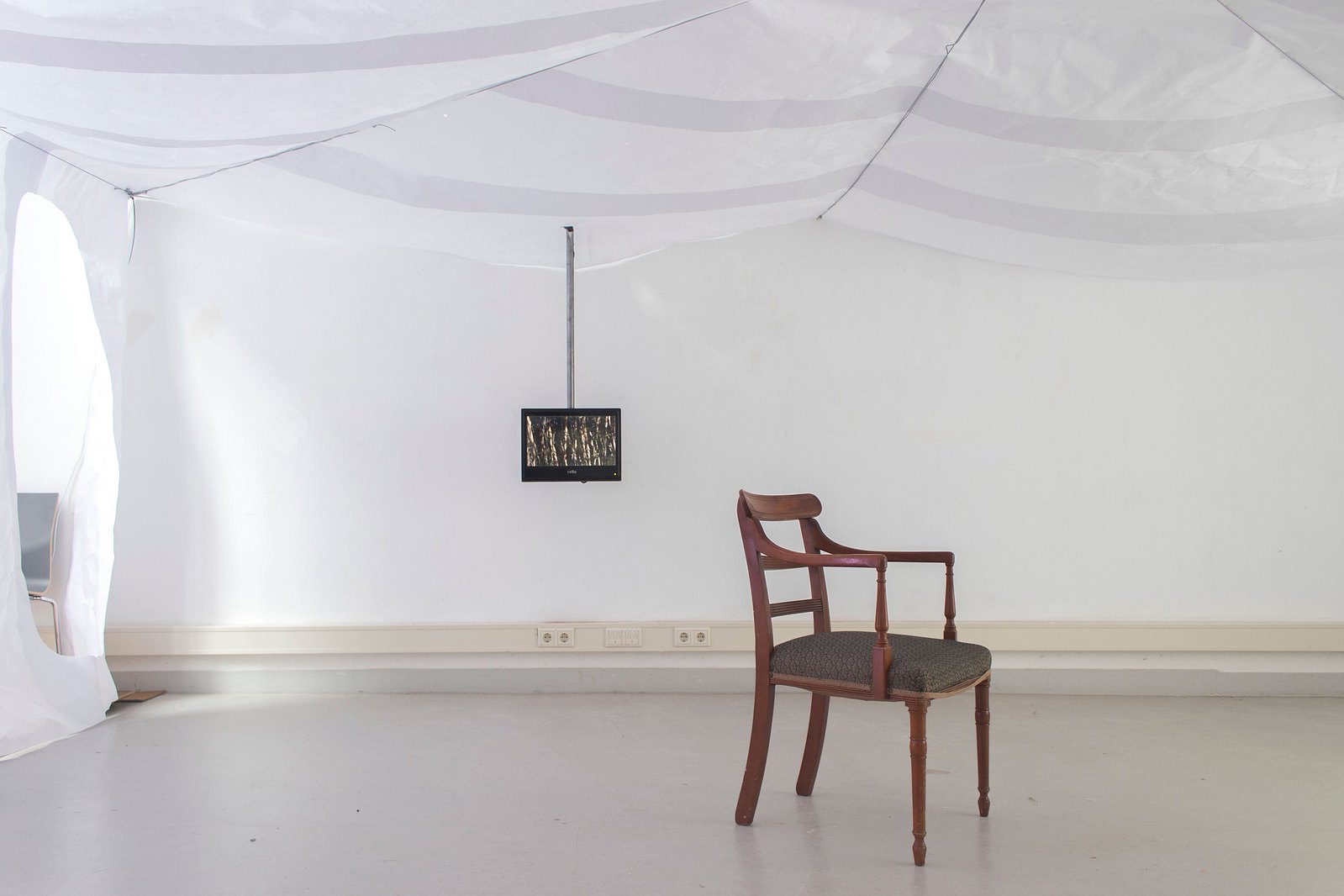 A room with paper ceiling and walls a wooden domestic chair. There is a video monitor on to behind coming down from the ceiling. Jamie Kane